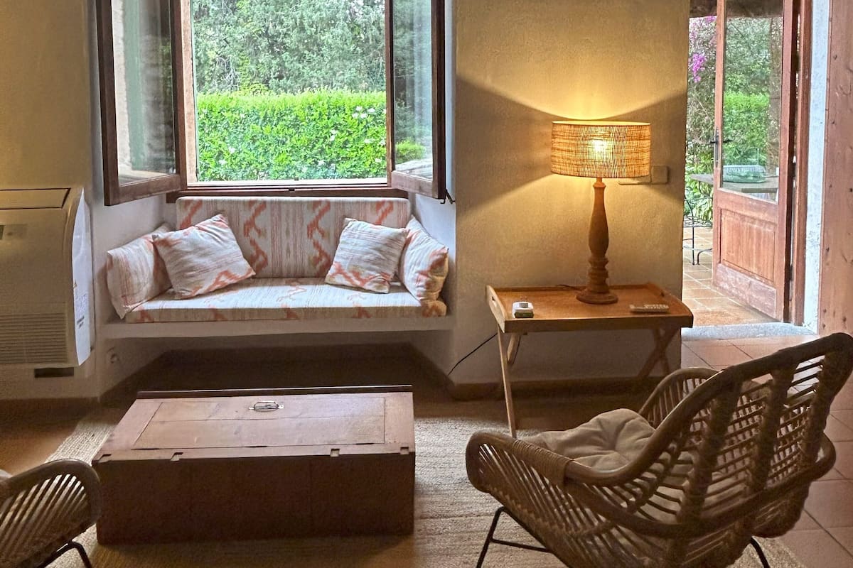 A cozy room with a built-in bench under a large window, a table with a lamp, and wicker chairs.