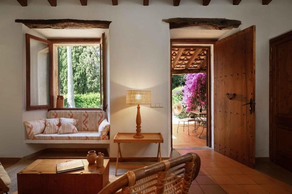A cozy room with a wooden bench, a table with a lamp, and a view of a garden through a window.