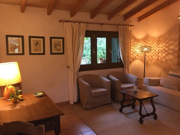 A cozy living room with a wooden table, two armchairs, a sofa, and a floor lamp. There are three framed pictures on the wall and a window with shutters.