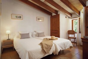 Una camera da letto con due letti, travi in legno al soffitto, una porta in legno, una sedia in vimini e un piccolo tavolo con una lampada.