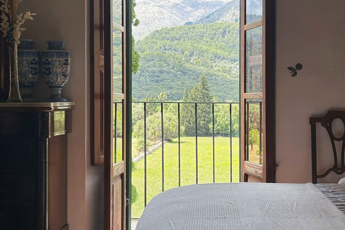 A bedroom with a large bed, a view of a green landscape through an open door, and a vase with flowers on a dresser.