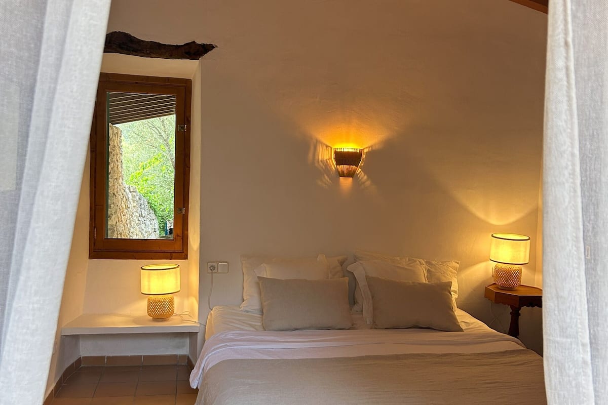 A bedroom with a bed, two bedside lamps, a window with a view of greenery, and a wooden ceiling beam.