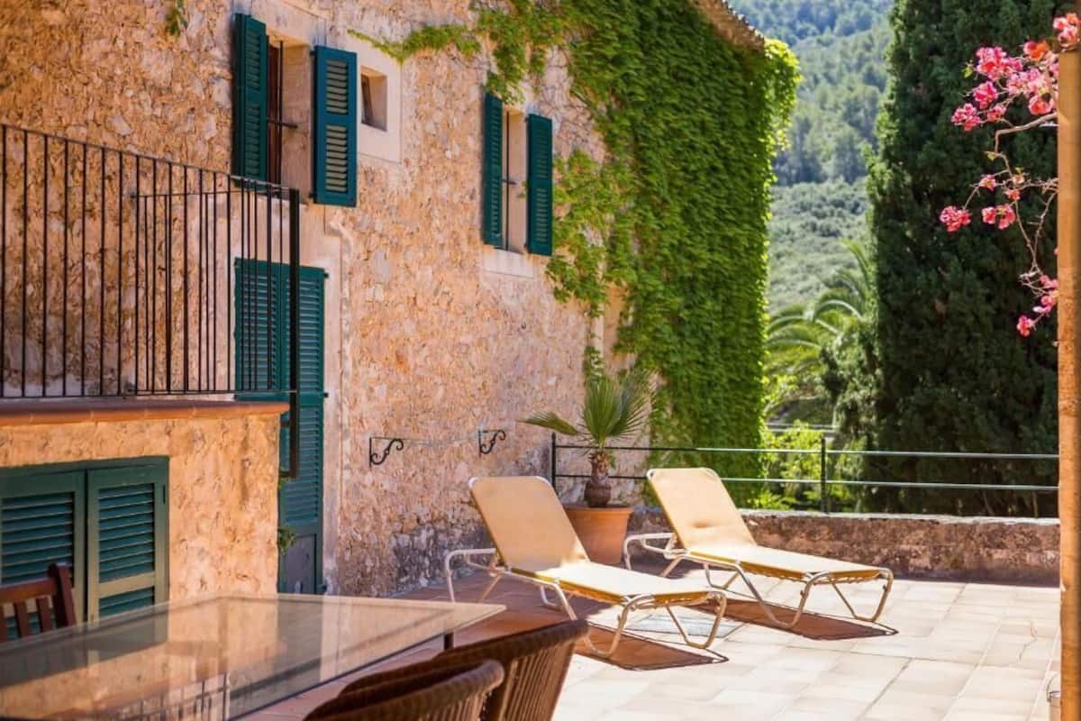 A stone building with green shutters, a balcony with two lounge chairs, and a potted plant.