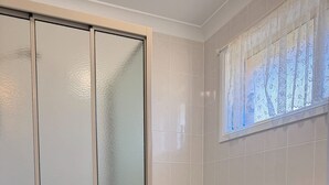 A bathroom with a toilet, a cabinet with drawers, a shower with frosted glass doors, and a window with lace curtains. - Waterfront Home with Bay Views | Sprindrift (Malua Bay)