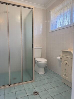 A bathroom with a toilet, a cabinet with drawers, a shower with frosted glass doors, and a window with lace curtains. - Waterfront Home with Bay Views | Sprindrift (Malua Bay)
