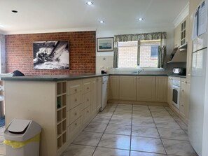 A kitchen with beige cabinets, a tiled floor, a trash bin, a window with curtains, and a picture on the wall.