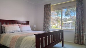 A bedroom with a wooden bed, patterned bedding, a bedside lamp, and a window with curtains. - Waterfront Home with Bay Views | Sprindrift (Malua Bay)