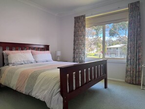 A bedroom with a wooden bed, patterned bedding, a bedside lamp, and a window with curtains. - Waterfront Home with Bay Views | Sprindrift (Malua Bay)