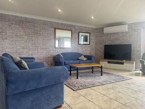 A living room with a brick wall, two blue sofas, a wooden coffee table, a flat-screen TV, and an air conditioning unit.