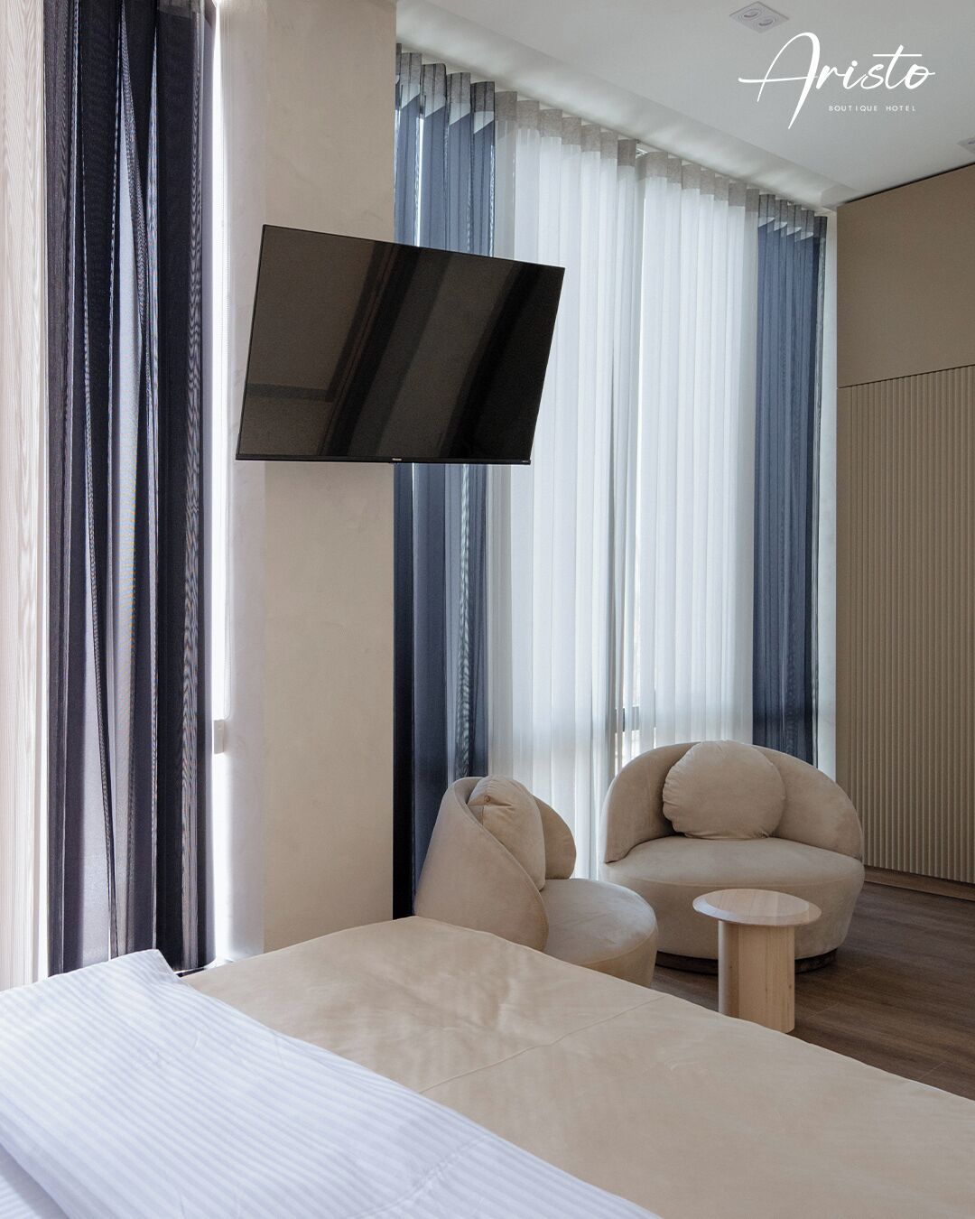 A modern hotel room with a bed, a sofa, a small round table, and a TV mounted on the wall.