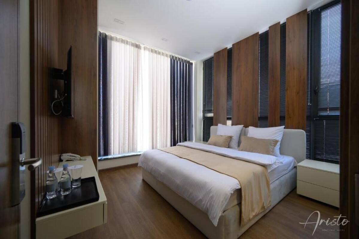 Ein modernes Hotelzimmer mit einem großen Bett, Holzpaneelen an den Wänden, einem Nachttisch mit Telefon und Wasserflaschen sowie großen Fenstern mit Jalousien.