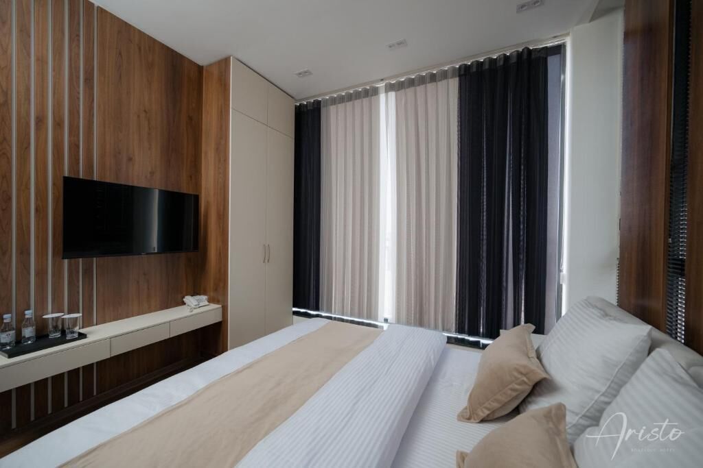Une chambre d’hôtel moderne dotée d’un grand lit, d’un téléviseur à écran plat fixé sur un mur en panneaux de bois, d’une table de chevet avec un téléphone et des lunettes, et d’une fenêtre avec des stores verticaux.