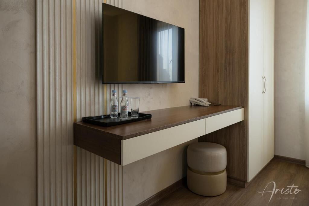 A modern hotel room with a flat-screen TV mounted on a wooden panel wall, a small desk with a telephone, and a cushioned stool.