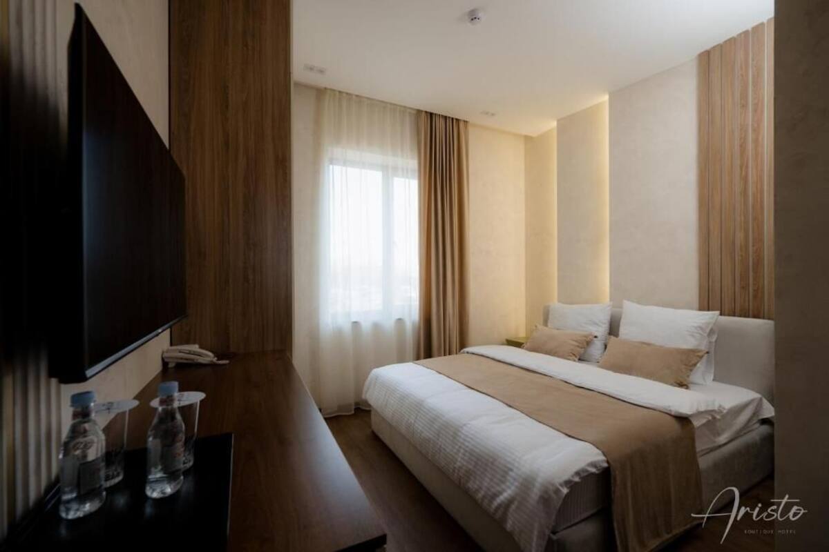 Ein Hotelzimmer mit Bett, Fernseher, Schreibtisch und Wasserflaschen sowie einem Fenster mit Vorhängen.