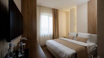 A hotel room with a bed, a TV, a desk with water bottles, and a window with curtains.