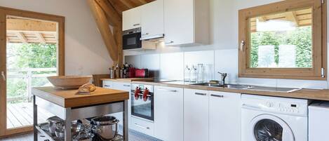 A modern kitchen with white cabinets, a built-in oven, a microwave, a sink, and a washing machine. There is a wooden table with a basket and a loaf of bread.
