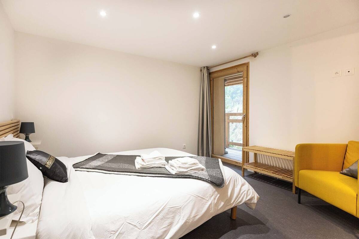 A hotel room with a bed, a bedside table with a lamp, a window with curtains, and a yellow armchair.