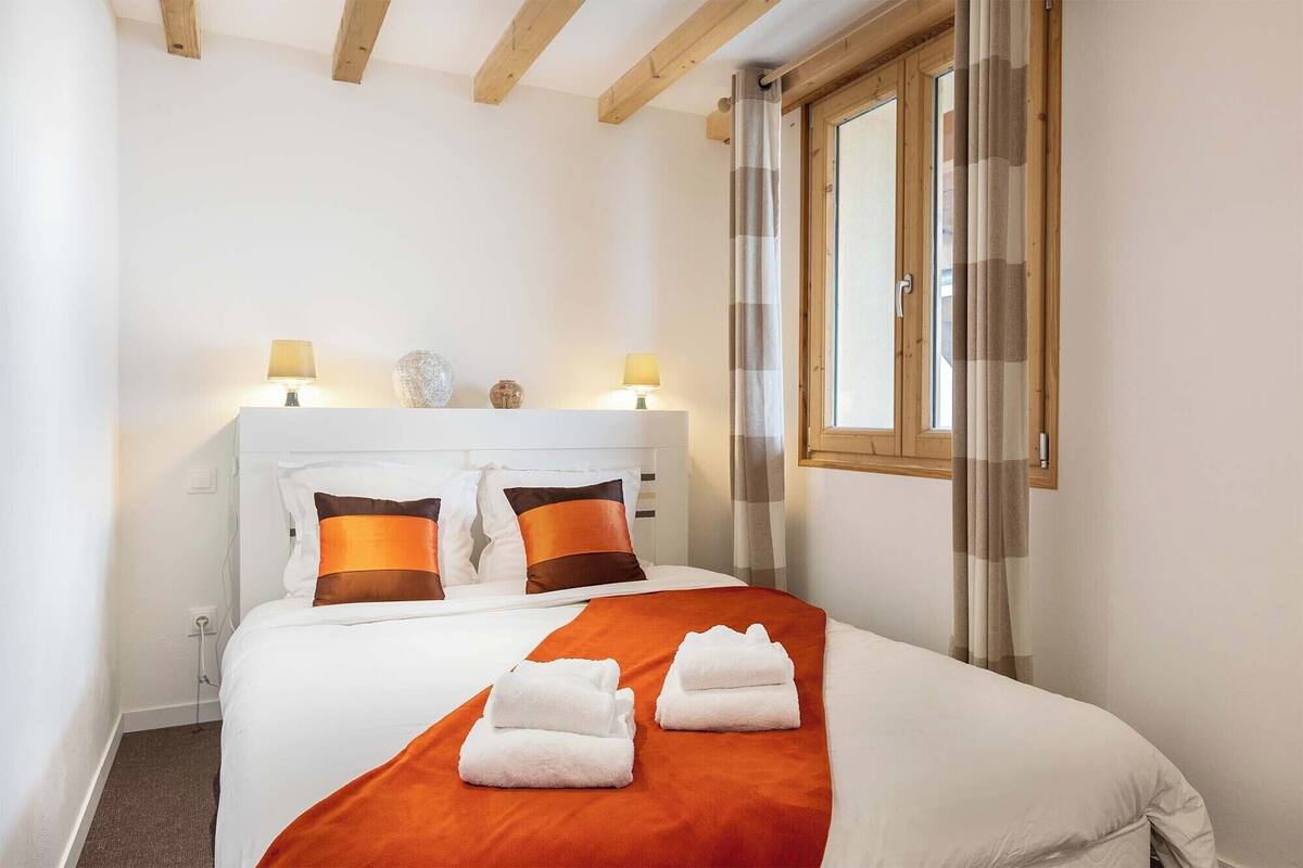 A neatly made bed with white and orange bedding, two bedside lamps, and a window with curtains.