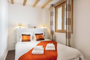Un lit bien fait, avec une literie blanche et orange, deux lampes de chevet et une fenêtre avec des rideaux.