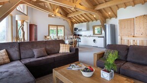 A spacious living area with a high ceiling and exposed wooden beams, a dark brown sofa, a wooden coffee table with magazines and a fruit bowl, and a view into the kitchen area. - Résidence Chalet Les Amis (Peisey-Nancroix)