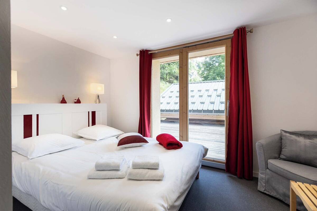A hotel room with a bed, two bedside tables, a red chair, and a view of the outside.