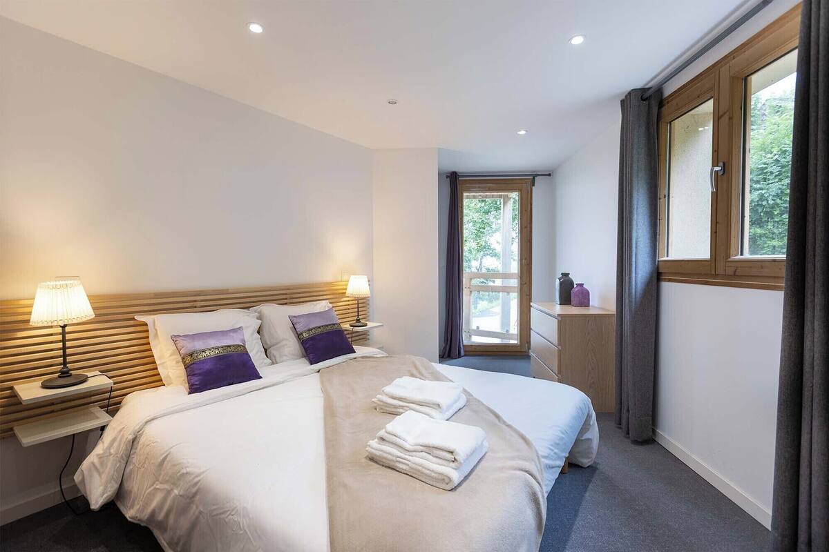 A modern hotel room with a large bed, bedside lamps, a wooden headboard, and a view through a window.