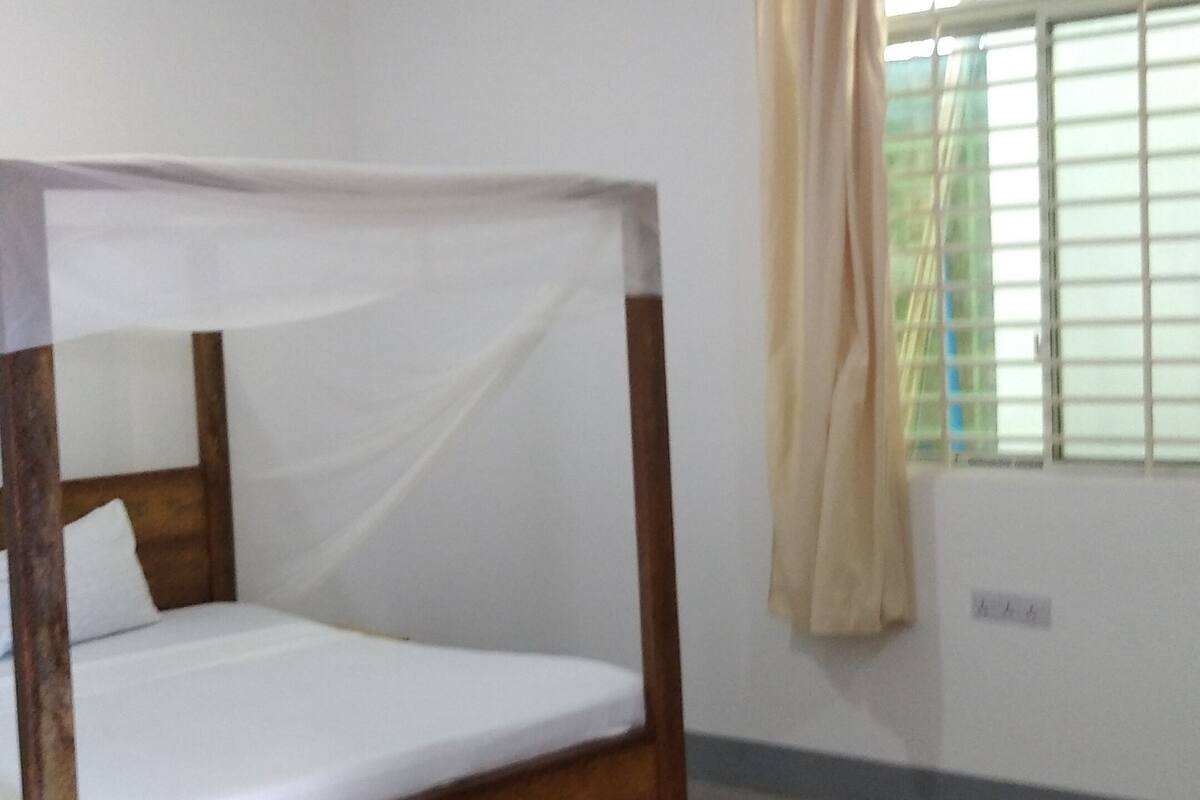 A simple, unadorned room with a single bed, a ceiling fan, and a window with curtains.