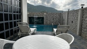 Un área de piscina con mesa y sillas, un muro de piedra y vistas a montañas.