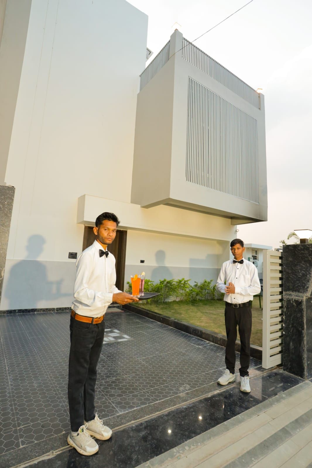 Two individuals standing outside a building with a modern design, a tiled floor, and a small garden area.