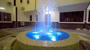 A fountain with water jets in a circular pool, surrounded by a tiled area with a low wall. - Vertex- View Hotel and Suites  (Awka)