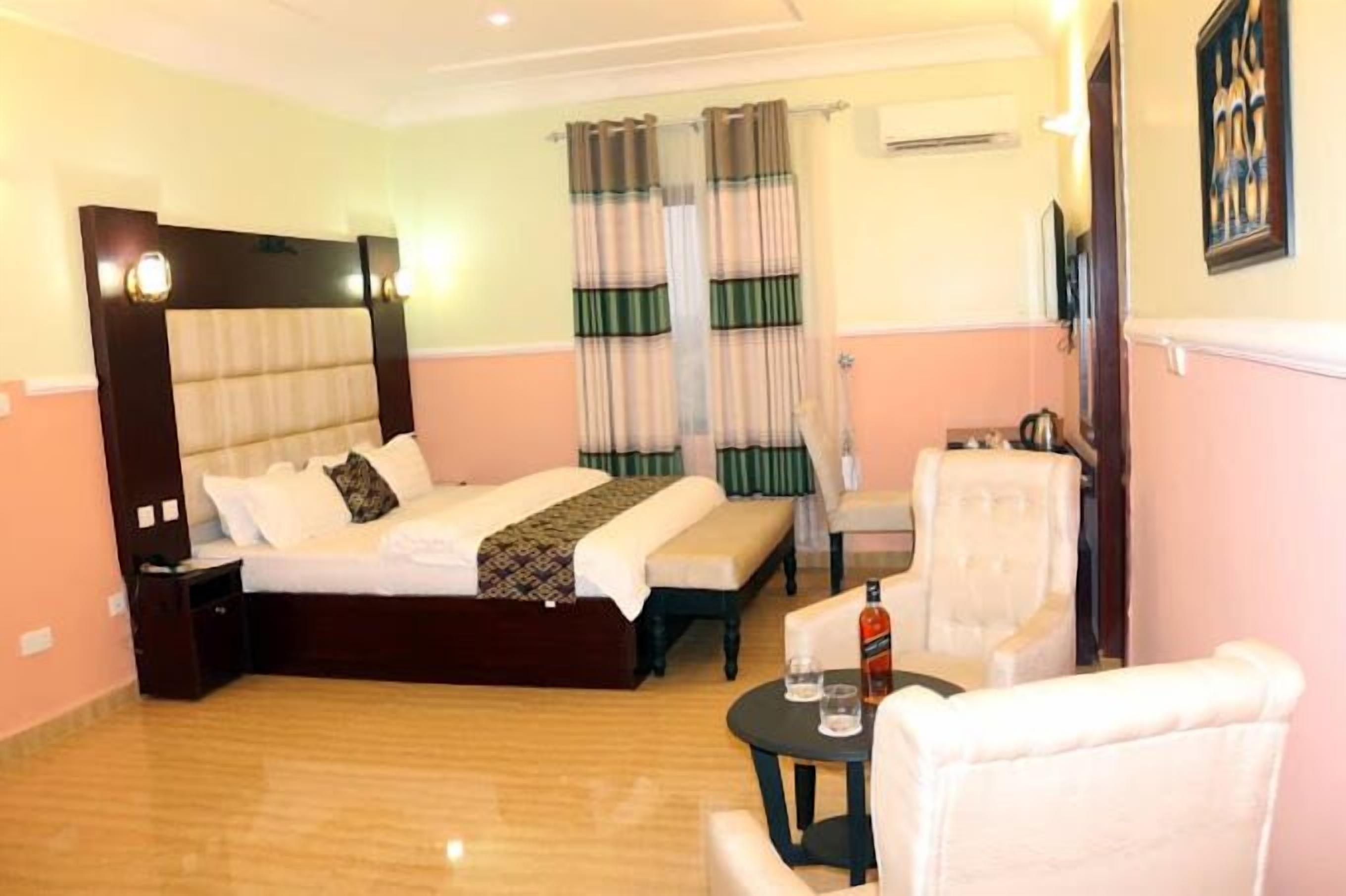 Quarto de hotel com uma cama grande, uma pequena mesa com uma garrafa, uma cadeira e uma janela com cortinas.