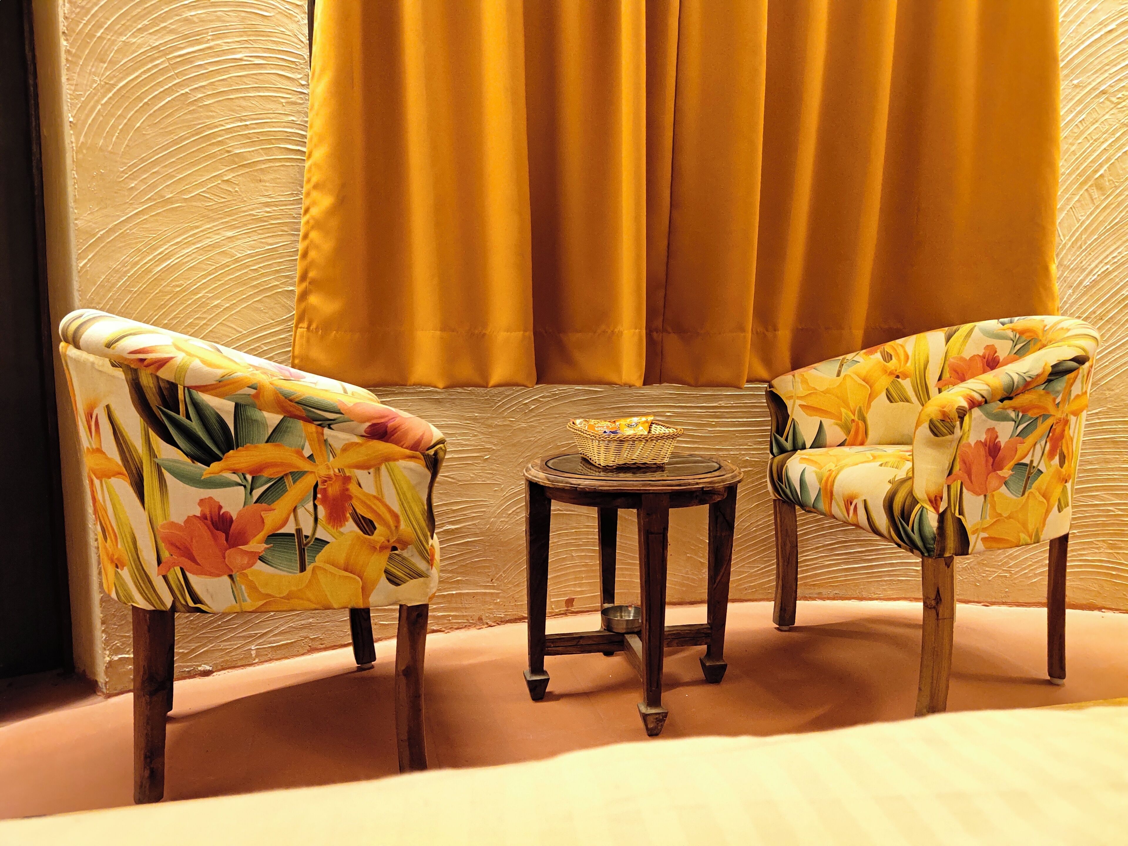 Duas poltronas florais, uma pequena mesa redonda com uma cesta e uma tigela, e uma janela com cortinas amarelas.