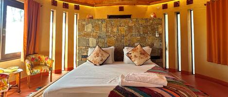 A room with a stone wall, a bed with pillows, a chair, and a window.