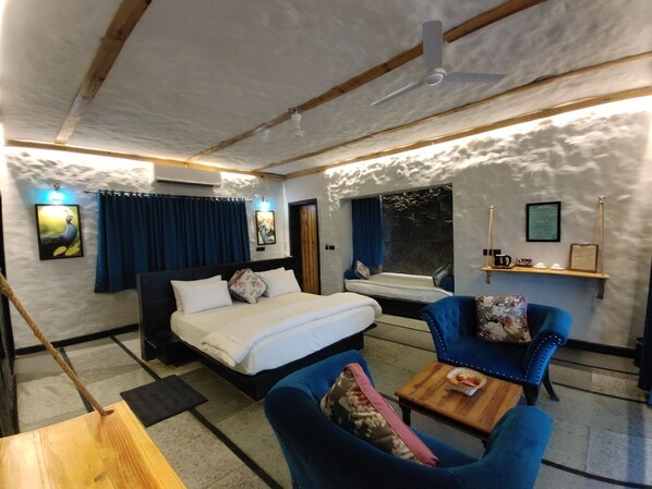 A bedroom with a bed, two blue armchairs, a wooden table, a window, and a ceiling fan. - Bella Basera (Kumbhalgarh)