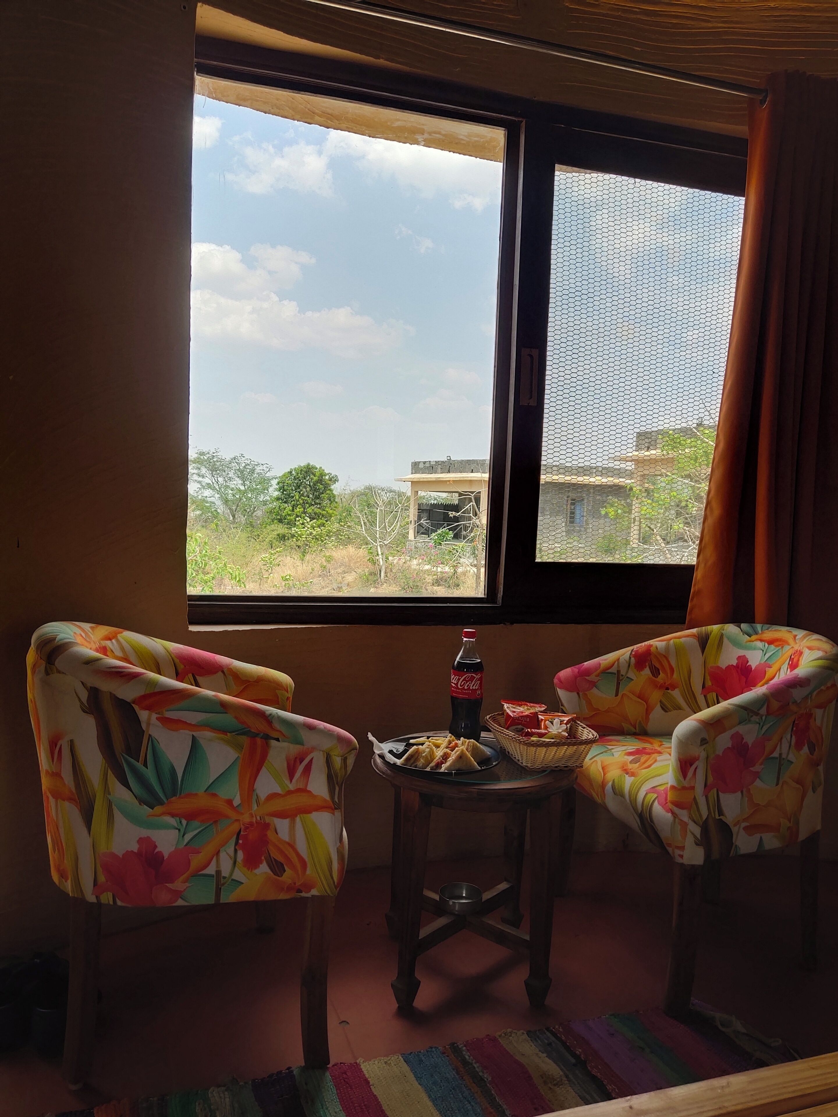 A room with two floral-patterned chairs, a small table with a bottle of Coca-Cola, a plate of food, and a colorful striped rug.