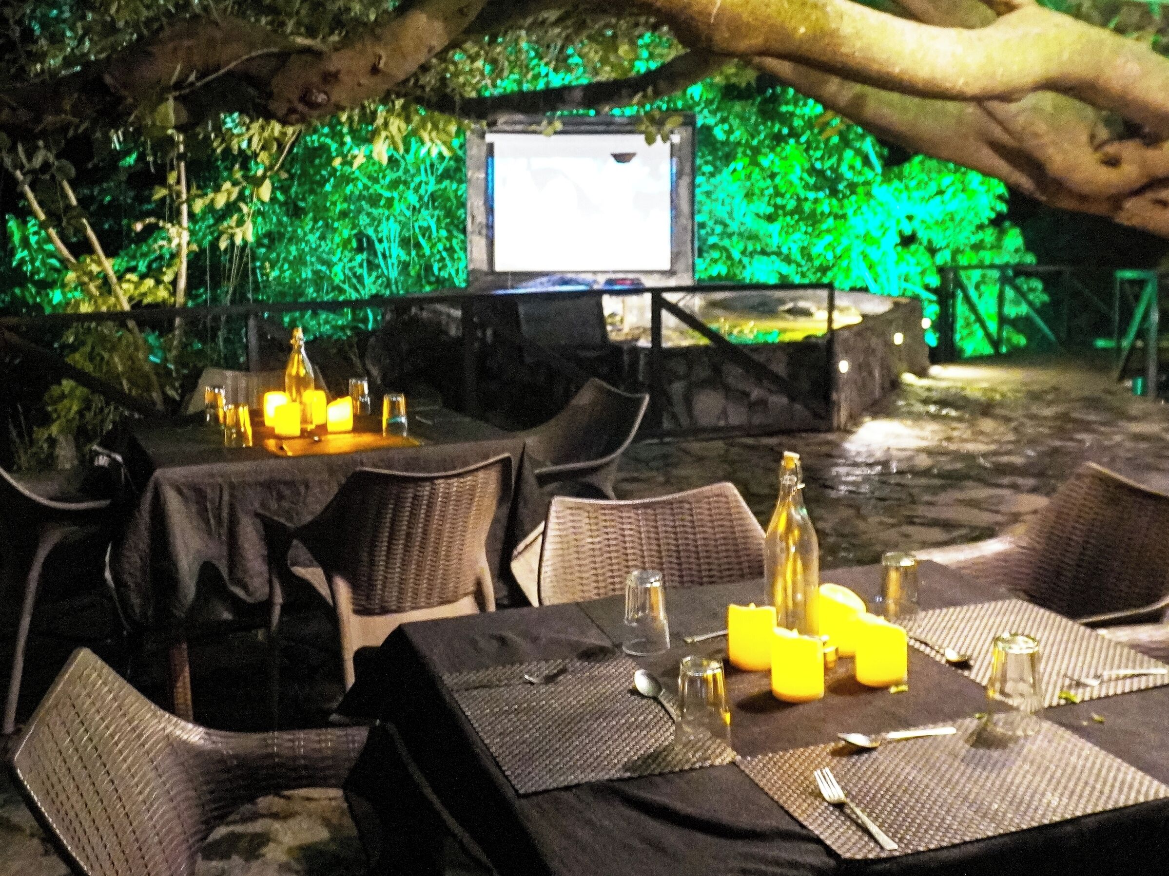 An outdoor dining area with tables set for two, covered with black tablecloths and equipped with candles, glasses, and cutlery.