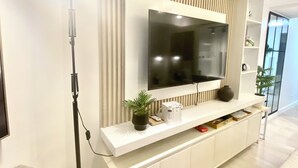A modern living room with a large flat-screen TV mounted on a wall with vertical paneling, a white console table with decorative items, a floor lamp, and a grey sofa. - Cozy studio in beautiful San Fernando with AC, WiFi and Pet Friendly  (San Fernando)