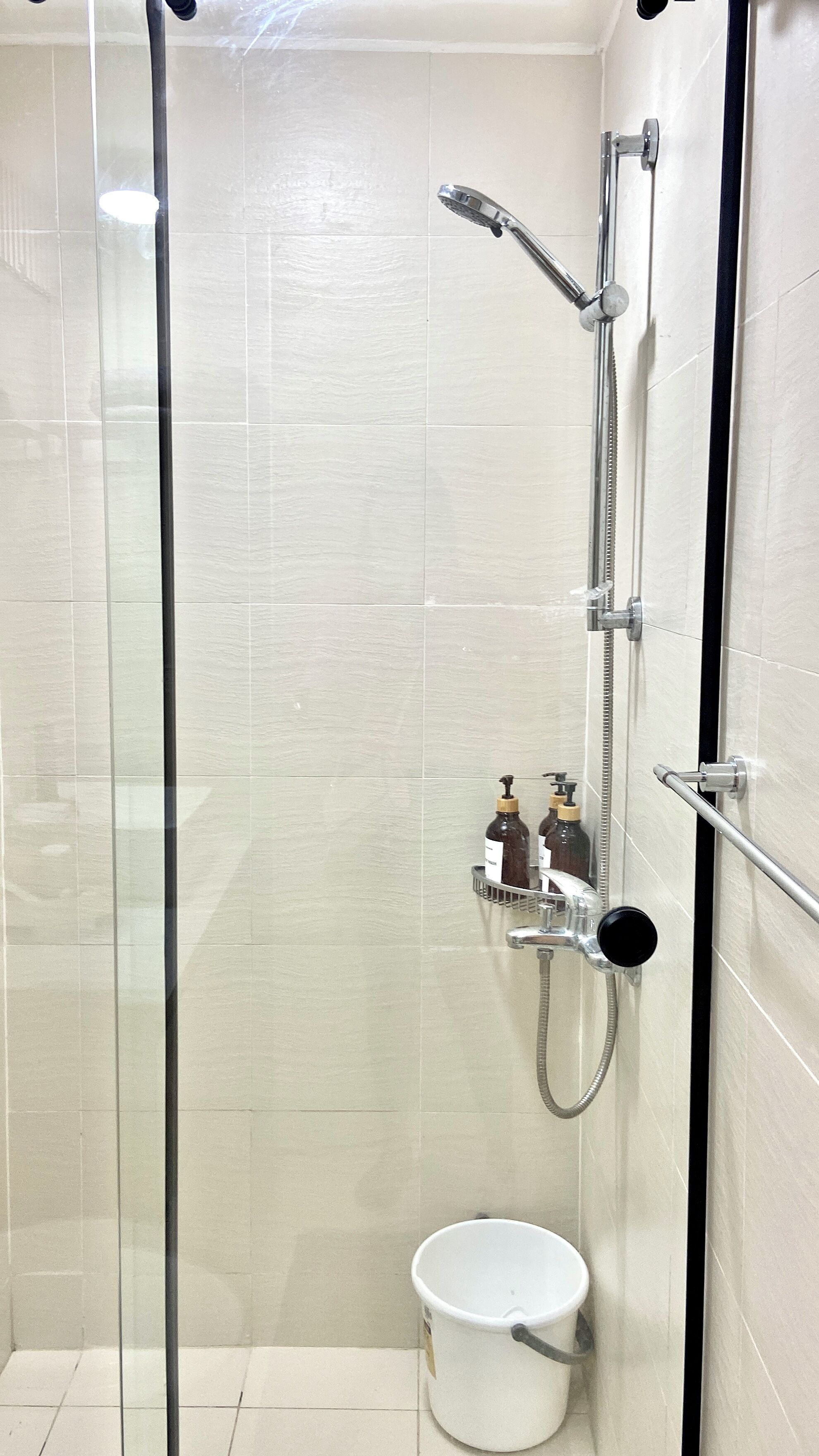A glass-enclosed shower stall with a handheld showerhead, a handheld shower attachment, and a white bucket.