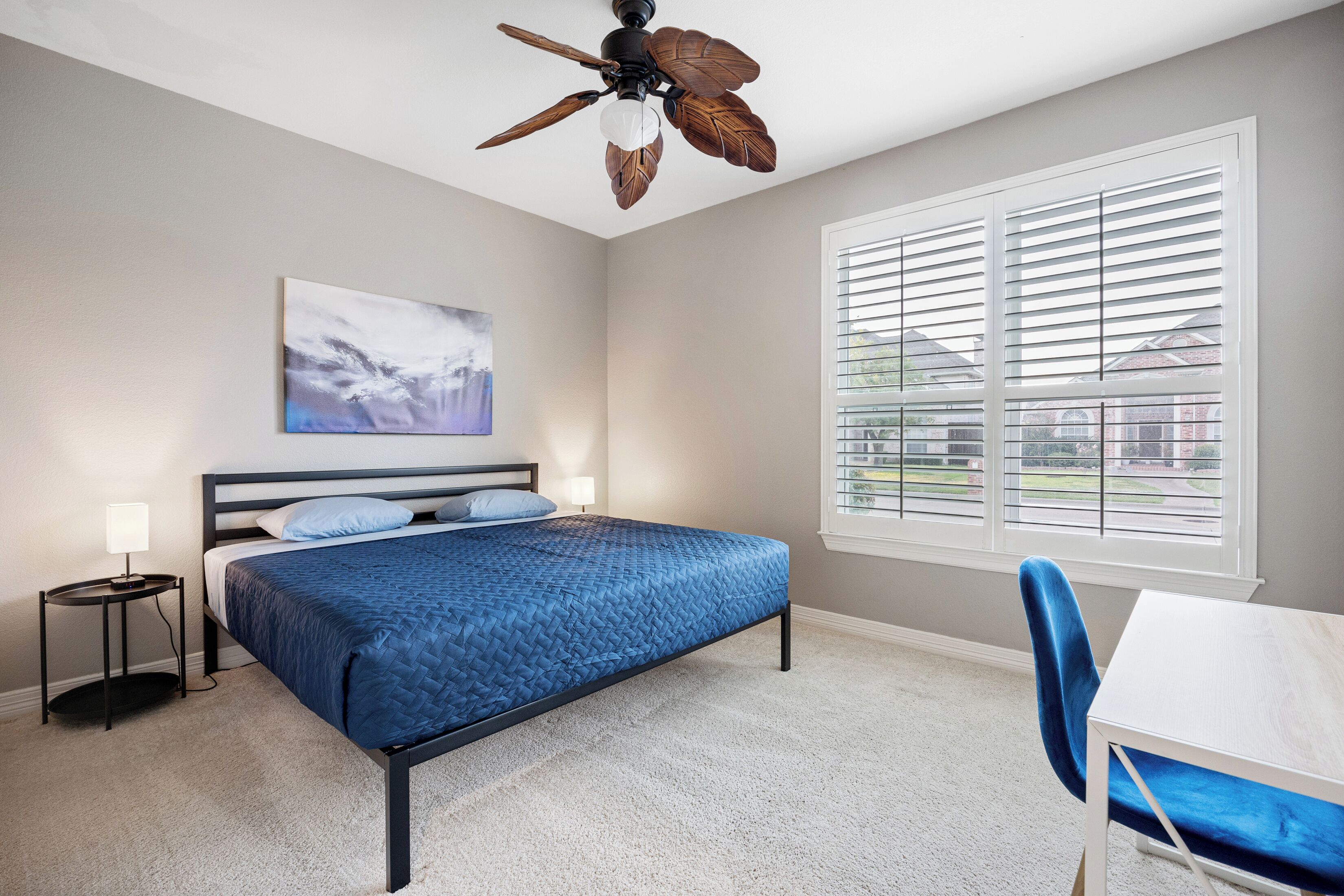 A bedroom with a bed, a ceiling fan, a window with blinds, a bedside table with a lamp, and a chair.
