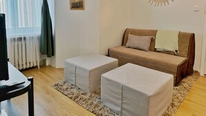 A living room with a sofa, two ottomans, a TV, a painting, and a sunburst mirror. - 1BR Demure Design Urban Living Space (Helsinki)