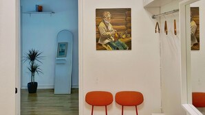 A room with a wooden floor, a potted plant, two orange chairs, a painting of a person playing a stringed instrument, and a ceiling with yellow and green drapes. - 1BR Demure Design Urban Living Space (Helsinki)