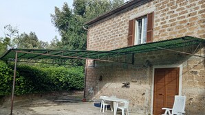 A rustic brick building with a wooden door and window shutters. There is a covered patio area with a table and two white plastic chairs.