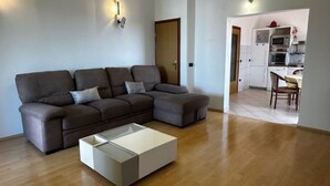 Un salon moderne avec un canapé d’angle, une table basse en verre et une cuisine en arrière-plan.
