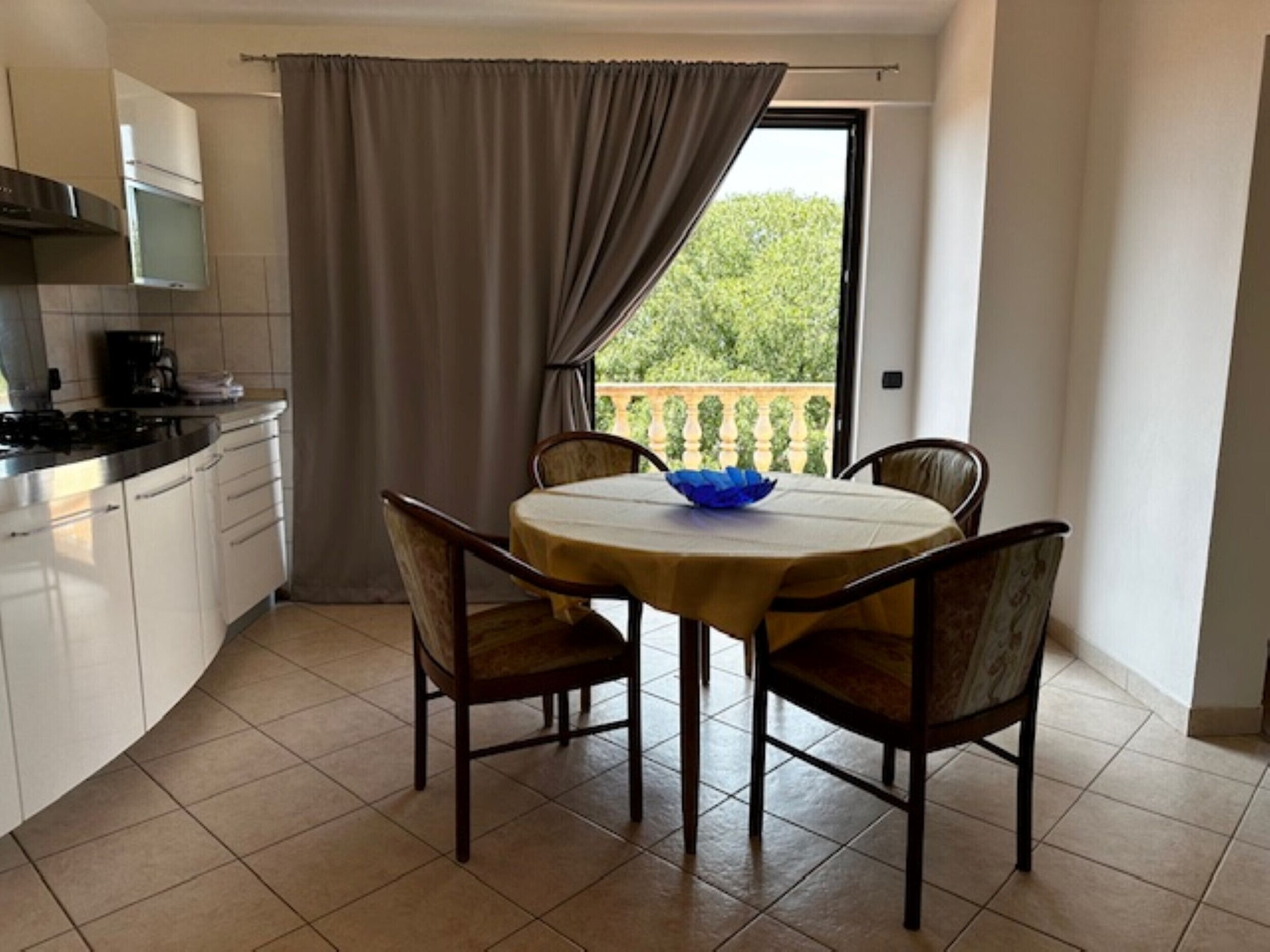 A kitchen with a dining table and chairs, a window with a view of greenery, and a curtain.