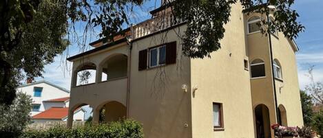 Ein zweistöckiges Gebäude mit einer beige farbenen Fassade, Rundbogenfenstern und einer Steinwand mit Blumentöpfen.
