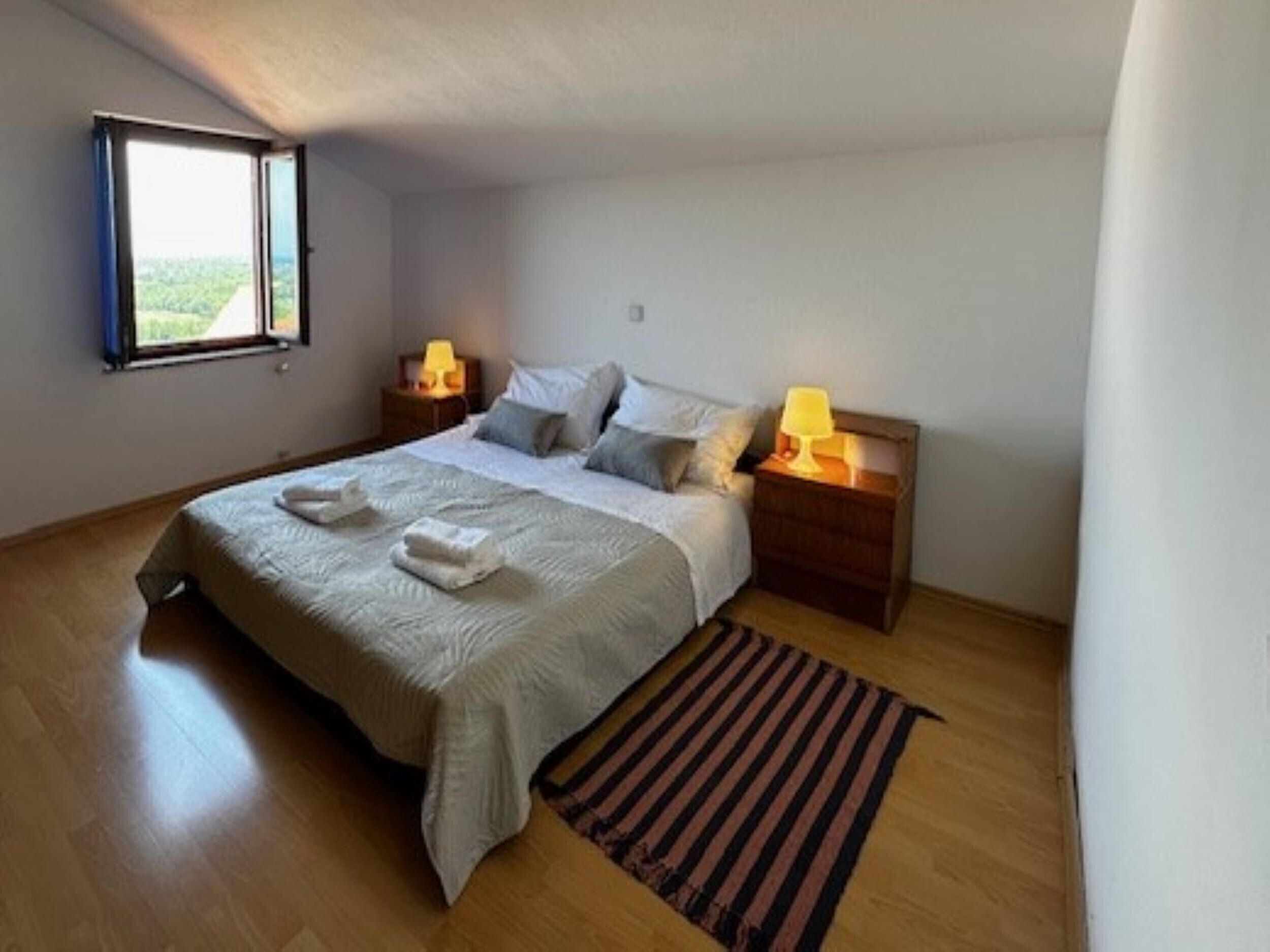 A bedroom with a bed, two bedside tables with lamps, a window, and a striped rug.