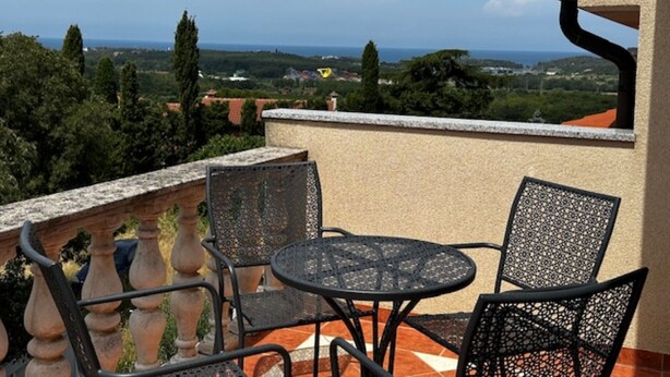 Un balcon avec une table et des chaises, offrant une vue panoramique sur un paysage verdoyant, avec des arbres et une vue lointaine sur la mer.