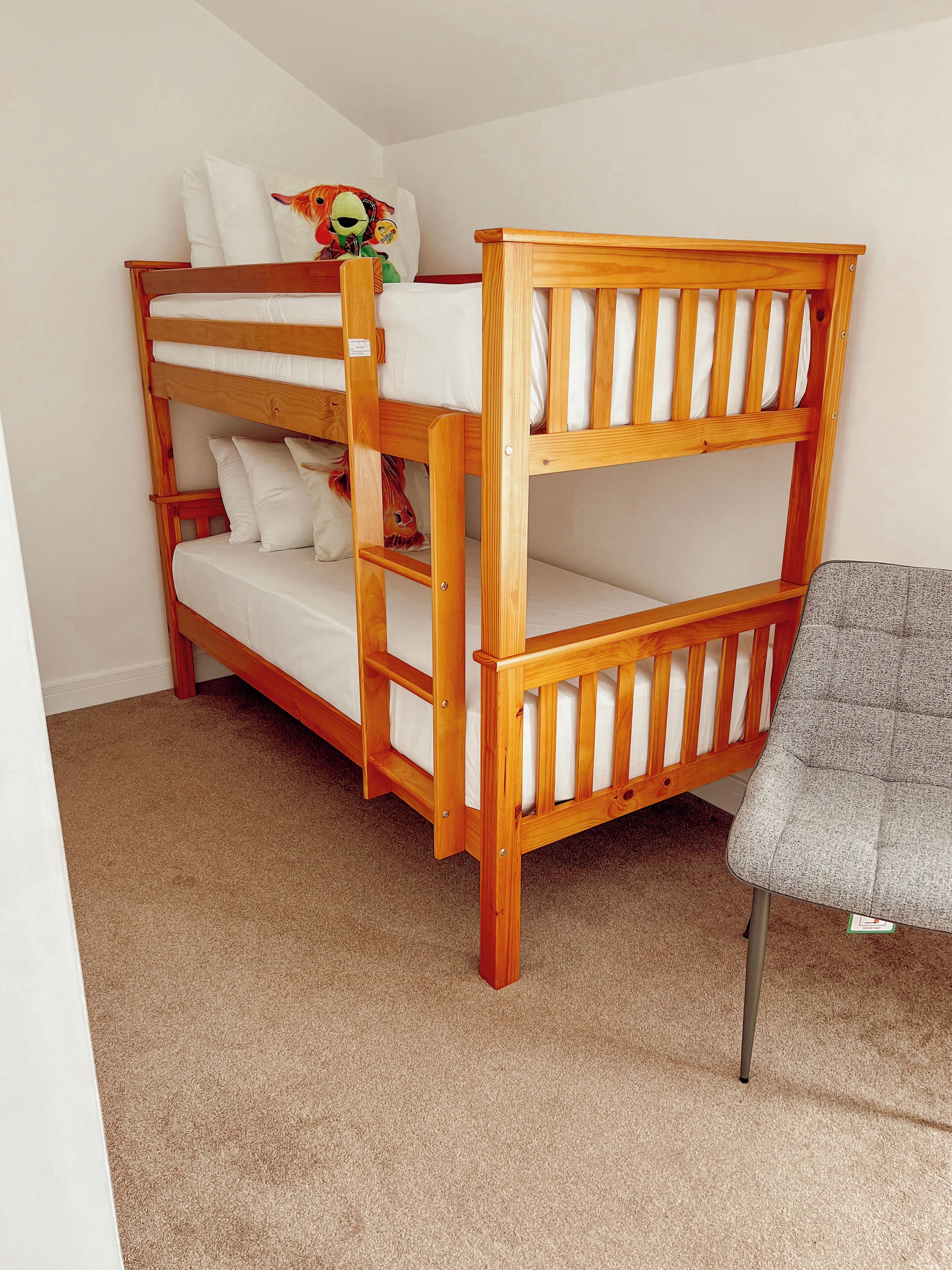 A bunk bed with a wooden frame and white bedding, a plush toy on the top bunk, and a gray chair in the corner.