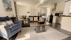 A spacious living room with a blue sofa, a coffee table, and a kitchen in the background. - Loch Ness Gate House Apartments (Fort Augustus)