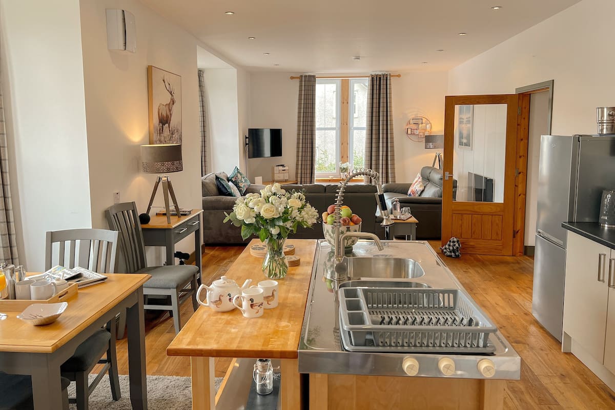 A modern kitchen with a central island, a dining area with a wooden table, and a living room with a sofa and TV.